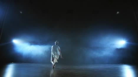 a-young-girl-in-a-white-dress-dances-contemporary-in-slow-motion-on-the-stage-with-smoke-in-spotlights.-Camera-stands-behind-her-filming-in-a-full-shot.